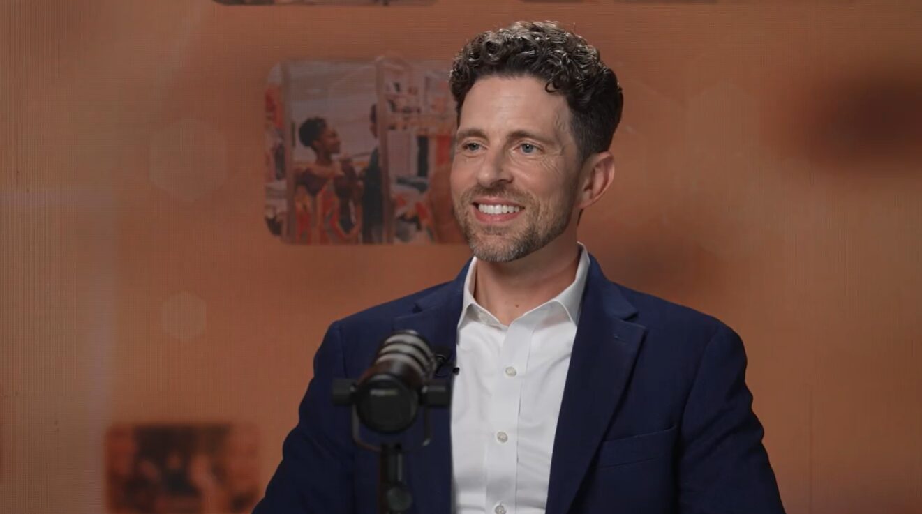 Smiling man in a navy blazer and white shirt speaks into a microphone in a recording studio.